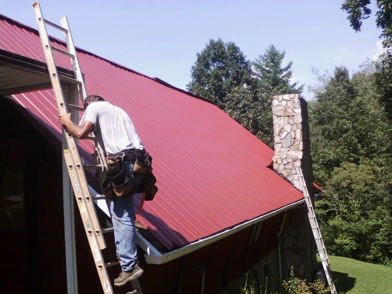 Roofer on a ladder beside a red metal roof and stone chimney — Soffit & Fascia Repair by Roofing Brothers in Keezletown